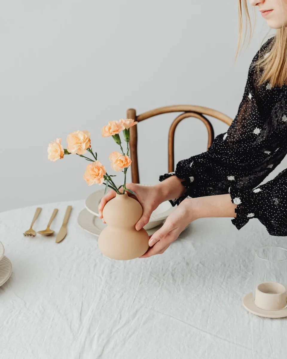 Hände stellen eine beigefarbene Keramikvase mit apricotfarbenen Nelken auf einen Tisch mit heller Leinendecke